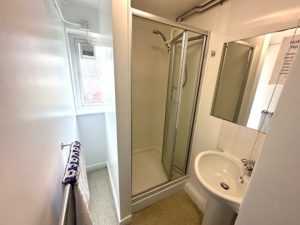 A narrow, compact bathroom with white walls and a light-coloured floor. On one side is a small frosted window that lets in natural light. Below it, a towel rail that holds a folded towel. Opposite the window is a corner shower enclosure with sliding glass doors and a mounted showerhead. Next to the shower is a small white sink with a curved basin and a wall-mounted mirror above it.