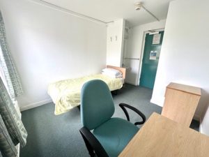 A small, simple bedroom with minimal furnishings. Against one wall is a single bed with a light yellow blanket and stacked pillows. In the foreground, there is a wooden desk with a green office chair. A small wooden cabinet stands to the side. The room has plain white walls, a carpeted floor, and a window with patterned curtains letting in natural light. At the back, a green door with posted notices leads to an adjoining area where a sink is partially visible.