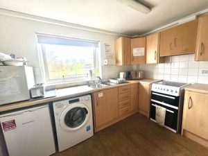 A small tidy kitchen with light wood cabinets and white tiles walls. Beneath a wide window is a countertop with stainless steel sink and drying rack. To the left is a compact refrigerator and washing machine under the counter. To the right is a free-standing cooker. Natural light streams in through the window, which looks onto a grassy area with trees. 